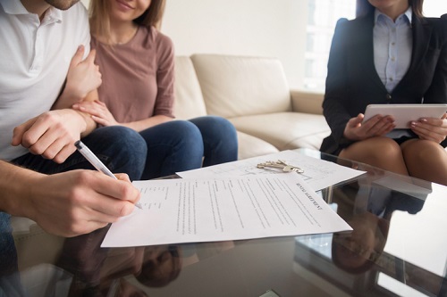 Signing rental agreement by young couple of renters or tenants, sitting on sofa with female real estate agent, male hand with a pen putting signature, long-term rent of property, close up